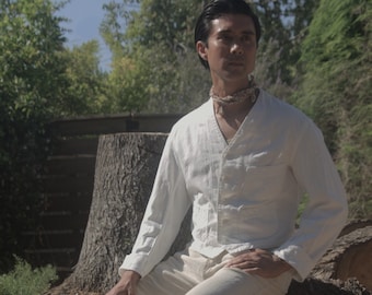 May include: A man wearing a white linen shirt and beige linen trousers sits on a tree stump. He is wearing a beaded necklace.