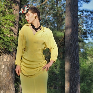 May include: A woman wearing a yellow dress with a floral print hat and a black beaded necklace. She is standing in a forest setting with trees and rocks in the background.