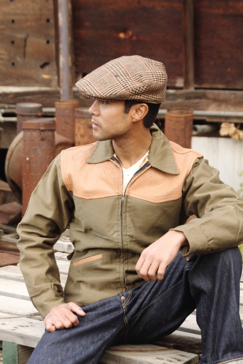 Pu&ograve; includere: Un uomo indossa un cappello piatto a quadri marroni e beige, una giacca verde con dettagli in pelle marrone e jeans blu. &Egrave; seduto su una piattaforma di legno.