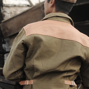 Pu&ograve; includere: Una giacca verde con una toppa di pelle marrone sul retro. La giacca ha una cintura di pelle marrone con una fibbia argentata. La persona che indossa la giacca indossa un berretto a quadri marroni e bianchi.