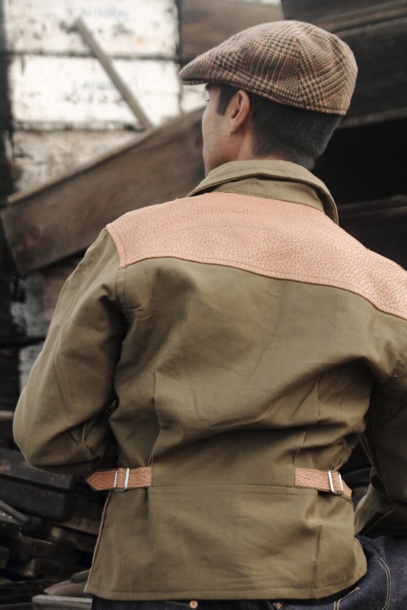 Pu&ograve; includere: Una giacca verde con una toppa di pelle marrone sul retro. La giacca ha una cintura di pelle marrone con una fibbia argentata. La persona che indossa la giacca indossa un berretto a quadri marroni e bianchi.