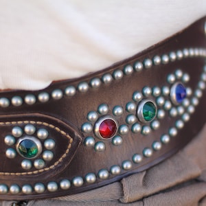 May include: A brown leather belt with silver studs and colorful gemstones. The belt has a decorative design with a row of silver studs and gemstones.