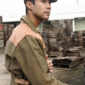 Pu&ograve; includere: Un uomo indossa un cappello da newsboy a quadri marroni e neri e una giacca utility verde con dettagli in pelle marrone. La giacca ha una cintura con una fibbia argentata.