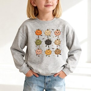 Puede incluir: Una sudadera gris claro de cuello redondo con un estampado gráfico de nueve calabazas de dibujos animados con varios patrones y colores, incluyendo naranja, verde, negro y rosa, cada una con brazos y piernas, y pequeñas estrellas y murciélagos.