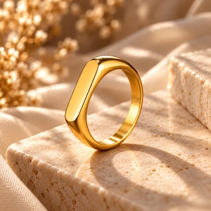 Peut inclure: Une bague en or poli dotée d'une face rectangulaire allongée et lisse ainsi qu'un anneau arrondi. La surface métallique présente une finition brillante et réfléchissante qui capte la lumière, offrant un aspect net et uni sans gravures.
