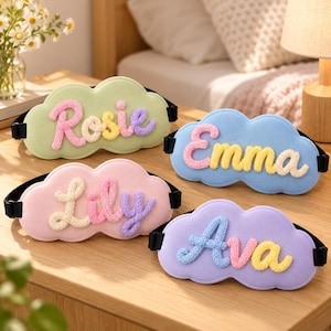 May include: Four cloud-shaped sleep masks with black elastic straps. Each mask features a soft pastel base colour and a name written in raised, multi-coloured knitted yarn letters. The names displayed are Rosie, Emma, Lily, and Ava in various colours.