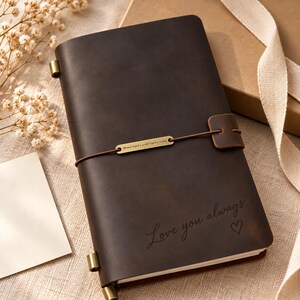 Könnte beinhalten: Ein dunkelbraunes Lederjournal mit der Gravur "Love you always" und einem kleinen Herzsymbol. Eine dünne braune Kordel umschließt den Einband und wird von einer rechteckigen Messingplatte mit der Gravur "Where there's a will there's a way" gehalten.