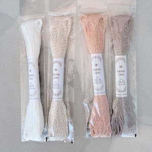 May include: Four skeins of Sashiko Lamé embroidery thread in individual clear plastic packaging. The colors from left to right are white, cream, light pink, and light gray. Each label indicates 40m, 96% cotton, 4% polyester, and made in Japan.