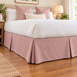 May include: A solid dusty rose bed skirt with a pleated design, featuring a textured fabric finish. The bed skirt is positioned beneath a white duvet, two dusty rose square pillows, and a rectangular cream-colored textured accent pillow.