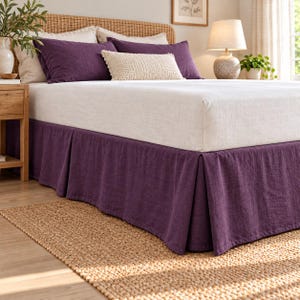 May include: A deep purple bed skirt with a pleated design, featuring a textured fabric finish. The bed skirt is positioned beneath a white mattress, two purple pillows, and a cream-colored rectangular accent pillow on a bed frame.