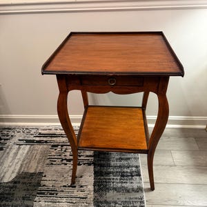 May include: A square wooden side table with a medium brown finish, featuring a single drawer with a small circular metal pull, a lower shelf, and four curved legs. The tabletop has a raised edge border, and the wood grain is visible.