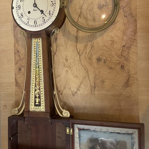 May include: A brown wooden banjo clock with a white face, black numerals, and the word Chelsea printed on the dial. The clock features a brass bezel, a decorative gold-toned panel with floral motifs, and a hinged lower door displaying a painted scene.