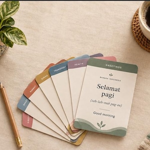 May include: A set of language learning flashcards fanned out, featuring the text Bahasa Indonesia, Selamat pagi, and Good morning. Each card has a colored header with labels like Greetings, Health, Transport, Dining, and Market, with a pencil nearby.
