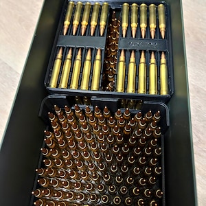 May include: A dark green metal ammunition box containing black plastic organisers filled with brass-cased rifle cartridges. The top section holds two rows of cartridges, while the bottom section holds a dense grid of vertically oriented rounds.