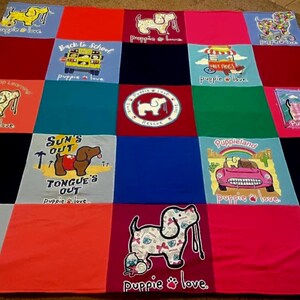May include: A patchwork quilt made from various colorful t-shirts featuring the Puppie Love brand logo and dog-themed graphics. The squares include illustrations of dogs, a school bus, a hot dog cart, and a car, with text like Sun's Out Tongue's Out.