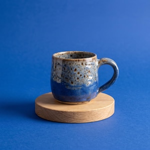 May include: A ceramic mug with a speckled finish featuring a gradient of light blue and brown at the top transitioning to a solid deep blue at the base, positioned on a circular light-colored wooden coaster with a visible natural grain pattern.