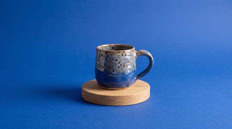 May include: A ceramic mug with a speckled finish featuring a gradient of light blue and brown at the top transitioning to a solid deep blue at the base, positioned on a circular light-colored wooden coaster with a visible natural grain pattern.