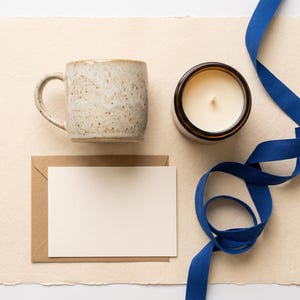May include: A speckled beige ceramic mug, a white candle in a dark brown glass jar, a blank cream-colored card, a brown paper envelope, and a long, winding royal blue ribbon arranged on a textured, rectangular, off-white paper sheet.