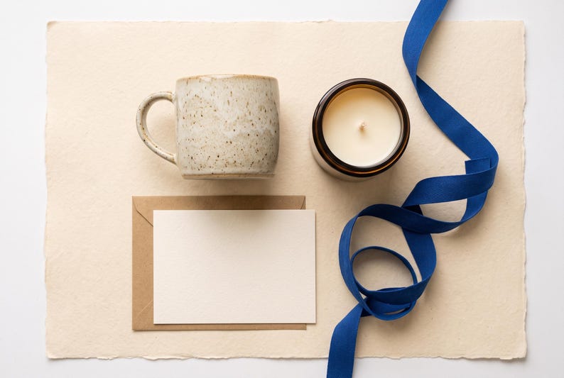 May include: A speckled beige ceramic mug, a white candle in a dark brown glass jar, a blank cream-colored card, a brown paper envelope, and a long, winding royal blue ribbon arranged on a textured, rectangular, off-white paper sheet.