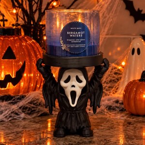 Puede incluir: Una vela azul de mezcla de cera de soja Bergamot Waters en un frasco de vidrio, sobre un portavelas negro con forma de figura con máscara de fantasma blanca y túnica negra. La etiqueta dice White Barn, Bergamot Waters, bergamot, blue waters, sandalwood.