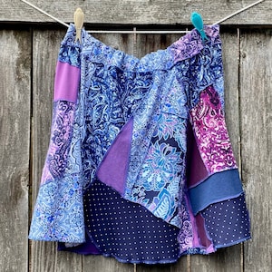 Puede incluir: Una falda de patchwork con una variedad de patrones florales y paisley en tonos de morado, azul y blanco. La falda tiene una cintura elástica y una silueta fluida.