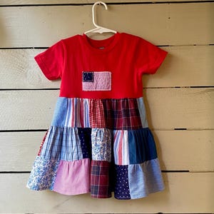 May include: A short-sleeved dress with a solid red bodice featuring a rectangular patch of a stylized flag. The skirt is constructed from multiple tiered panels of fabric in various blue, red, and white patterns, including plaid, stripes, and floral prints.