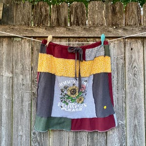 May include: A patchwork skirt with a variety of fabrics in shades of yellow, gray, burgundy, and green. The skirt features a drawstring waist and a central panel with the text "Honey and Flowers Please" surrounding a floral design.