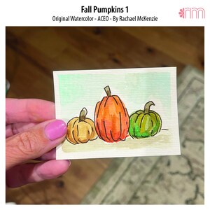 Könnte beinhalten: Ein kleines rechteckiges Aquarellbild mit dem Titel Fall Pumpkins 1 von Rachael McKenzie, das drei Kürbisse in Gelb, Orange und Grün mit schwarzen Tuscheumrissen vor einem hellgrünen Hintergrund auf strukturiertem Papier zeigt.