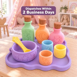 Peut inclure: Un ensemble de jouets en silicone sur un plateau violet en forme de nuage, comprenant un bol violet avec une cuillère jaune, trois flacons à bouchons marron et trois petits gobelets bleu, jaune et orange, ornés de motifs célestes et floraux.