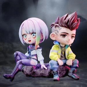 May include: Two stylized chibi-style figurines sitting side-by-side. The left figure has short, multi-colored hair, a white jacket, and purple pants. The right figure has spiky brown hair, a yellow jacket, a gold cross necklace, and blue pants.