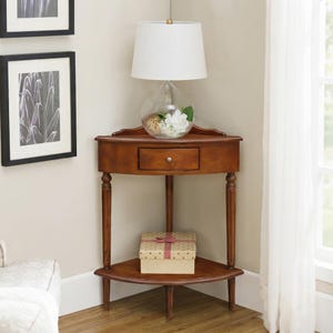 May include: A brown wooden corner table featuring a single drawer with a small round knob, three slender legs, and a lower triangular shelf. A glass lamp with a white shade and a decorative box are placed on the table surfaces.