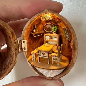 Pode incluir: Um diorama em miniatura dentro de uma casca de noz oca, apresentando um pequeno conjunto de jantar de madeira com mesa, banco e cadeira. O interior é iluminado por uma pequena luz e inclui uma prateleira com roupa de cama dobrada, uma planta e arte de parede.