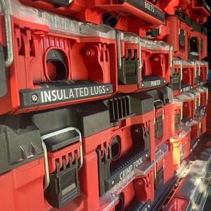 Op de afbeelding: Een stapel rode en zwarte modulaire gereedschapsorganizers met doorzichtige deksels en zwarte sluitingen. Elke unit heeft een zwart label met witte tekst, waaronder Insulated Lugs, Bolts, Printer, Crimp Tool en Switch Board in een verticaal systeem.