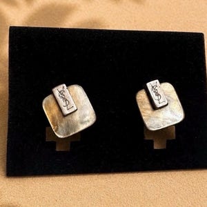 May include: A pair of silver-toned square earrings featuring a brushed metallic finish and a raised rectangular tab at the top. Each tab is engraved with the YSL logo. The earrings are mounted on a rectangular black display card.