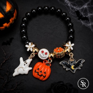 Op de afbeelding: Een elastische armband met glanzende zwarte kralen en goudkleurige tussenstukjes. De armband bevat vijf bedels: een wit spookje, een lachende oranje pompoen, een witte schedel met rode ogen, een oranje bloem en een zwarte vleermuis met een gele maan.