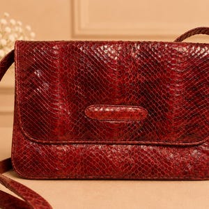 Pode incluir: Uma bolsa de ombro retangular feita de couro de cobra vermelho escuro com um padrão de escamas distinto. A bolsa apresenta um fecho de aba frontal com um detalhe de aba horizontal e uma alça de ombro fina a condizer, com acabamento brilhante.