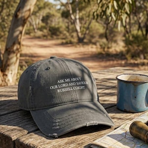 May include: A charcoal grey baseball cap with a distressed brim and white embroidered text that reads: ASK ME ABOUT OUR LORD AND SAVIOUR RUSSELL COIGHT. The cap features a structured crown with ventilation eyelets and a curved visor.