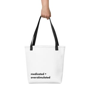 May include: A white tote bag with black handles featuring the black text "medicated + overstimulated" printed in a sans-serif font on the lower left side. The bag has a rectangular shape with a flat bottom and two sturdy shoulder straps.