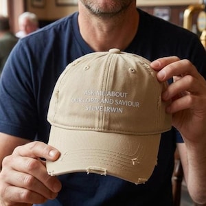 May include: A beige baseball cap with a distressed, worn-in finish and frayed edges. The front of the cap features white embroidered text that reads: ASK ME ABOUT OUR LORD AND SAVIOUR STEVE IRWIN, centred on the crown above the curved brim.
