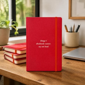 May include: A bright red hardcover notebook with an orange elastic closure band. The front cover features the white printed text: things I absolutely cannot say out loud. The notebook is positioned vertically, with three additional identical notebooks stacked.