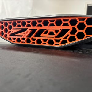 May include: A gray automotive grille insert featuring a bright orange honeycomb pattern and the black text Z10. The component has a sleek, tapered frame and a textured finish, designed to fit into a vehicle front end with a geometric design.