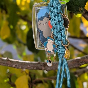 May include: A teal paracord keyring featuring a rectangular frame with a Snorlax illustration, a small Snorlax enamel charm, a Poké Ball charm, a banana charm, and a green flower bead, all attached to a gold-toned metal lobster clasp.