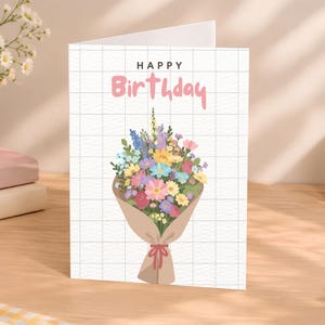 May include: A white greeting card with a black grid pattern featuring the text HAPPY in black and Birthday in pink script. The centre displays a colourful bouquet of flowers wrapped in brown paper with a tied red ribbon at the base.