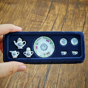 May include: A miniature porcelain tea set featuring a white teapot, creamer, sugar bowl, large plate, two small saucers, and two tiny teacups, all decorated with delicate pink and green floral patterns, presented in a dark blue velvet tray.