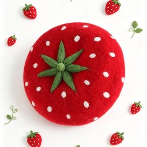 May include: A large, round, red felted wool pincushion shaped like a strawberry with white embroidered dots and a green felted stem. It is surrounded by several smaller felted strawberry embellishments and green leaf accents.