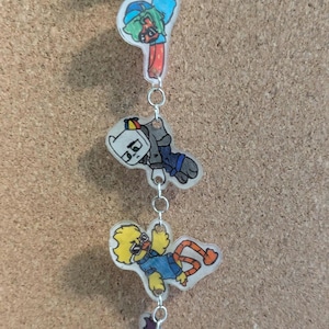 Könnte beinhalten: Ein vertikaler Hängeanhänger mit vier verschiedenen Acrylfiguren im Cartoon-Stil, die durch silberfarbene Metallringe verbunden sind. Die Figuren zeigen einen Charakter mit grünen Haaren, einen weißköpfigen Charakter, eine gelbe vogelartige Figur und eine dunkle Figur.