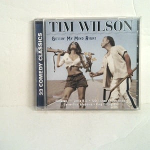 May include: A Tim Wilson Gettin' My Mind Right CD case featuring a cover image of a person in a white shirt and shorts holding a rifle and another person in a light-colored outfit holding a shovel on a road. The text 33 Comedy Classics is visible.