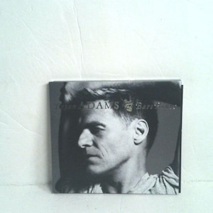 May include: A square music CD case featuring a black and white portrait of a person with their arm raised behind their head. The text Bryan Adams Bare Bones is printed across the center in a dark font, with a small skull icon between the words.