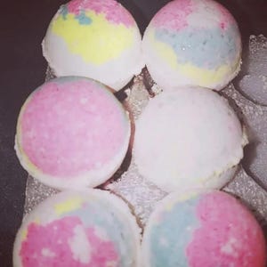 May include: Six spherical bath bombs arranged in two columns of three. Each bath bomb features a unique blend of pastel colours including pink, yellow, blue, and white, with a textured, porous surface characteristic of compressed bath fizzies.