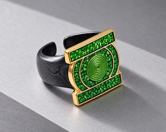 Puede incluir: Un anillo con una banda negra mate de superficie texturizada y un emblema rectangular en tono dorado. El emblema muestra un fondo verde brillante y texturizado con un motivo circular central compuesto por anillos verdes concéntricos.